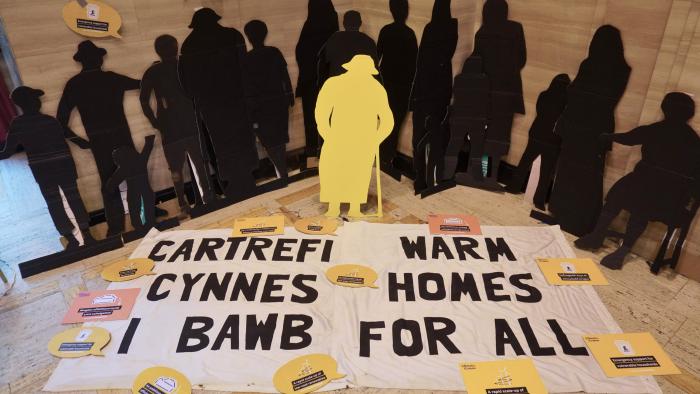 Black silhouettes behind a sign that says 'Warm Homes for all' in English and Welsh/Silwetau du tu ôl i arwydd sy'n dweud 'Cartrefi Cynnes i Bawb' yn Gymraeg a Saesneg